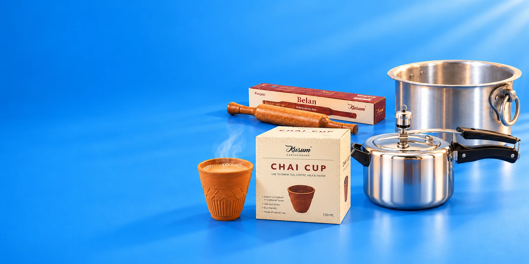 Kusum aluminium cooking pot, pressure cooker, steaming chai cup and Punjabi belan with retail cartons on a sunlit blue backdrop